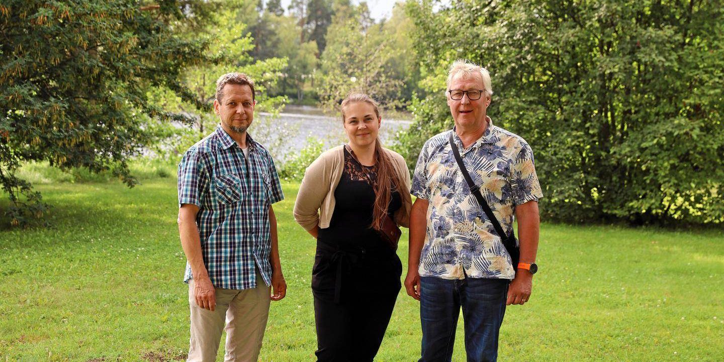 Kolme henkilöä seisoo nurmikolla ja taustalla on järvi ja puita. Vasemmalla Kai Munukka, keskellä Katja Kalenius ja oikealla Erkki Lehtimäki.