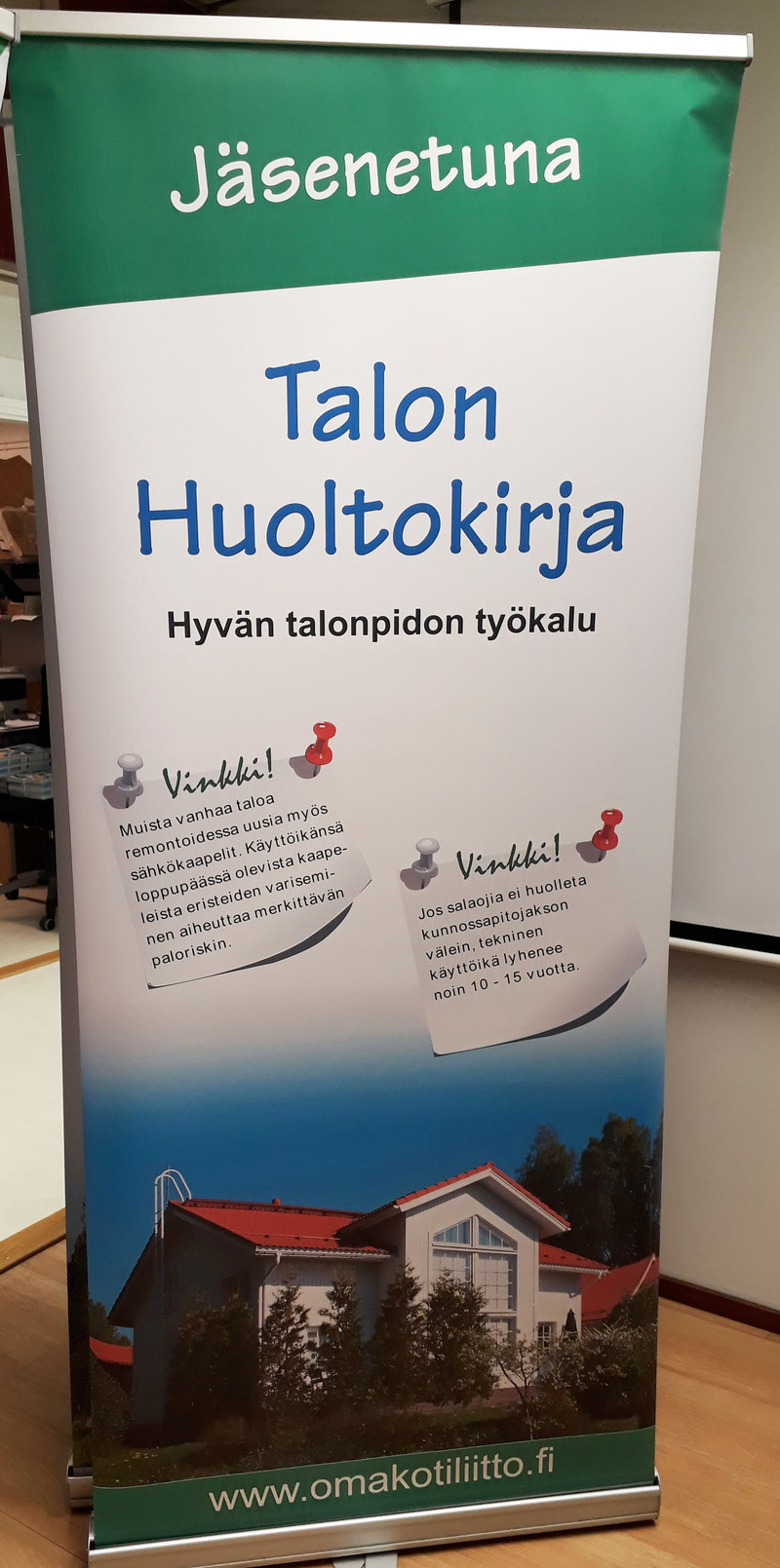 Omakotiliiton roll-up, jossa on esitelty jäsenetuna saatavaa talon huoltokirjaa.