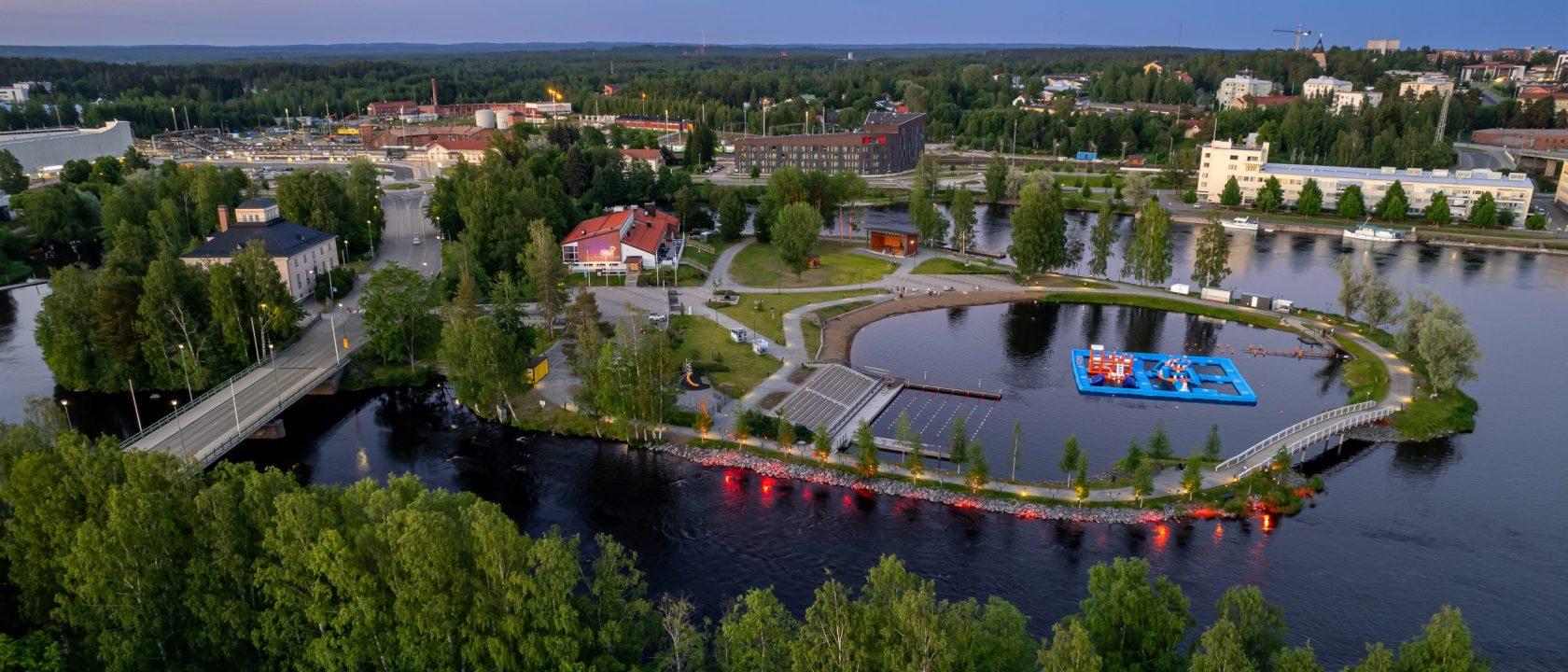 Ilmakuva Joensuusta, etualalla näkyy vettä ja puistomaista maisemaa, taustalla rakennuksia. Ilta on hämärtynyt, ja katulamppujen valo heijastuu joen pinnasta.