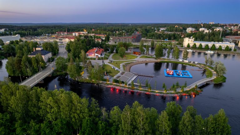 Ilmakuva Joensuusta, etualalla näkyy vettä ja puistomaista maisemaa, taustalla rakennuksia. Ilta on hämärtynyt, ja katulamppujen valo heijastuu joen pinnasta.