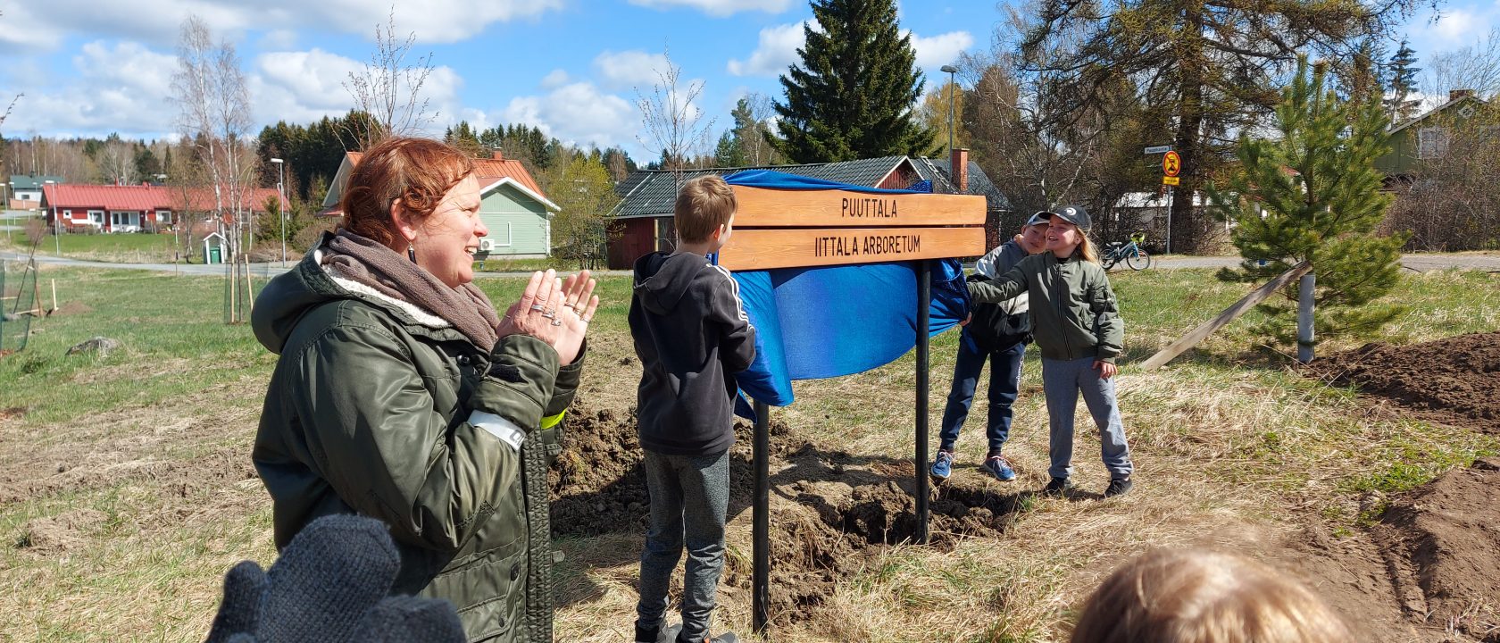 Puuttalan nimikyltin paljastetaan. Kolme koululaista on nostanut sinisen kankaan nimikyltin yläpuolelta ja yleisö taputtaa.