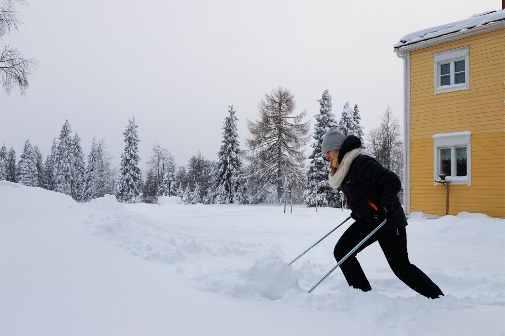 Nainen kolaa lunta keltaisen puutalon pihalla