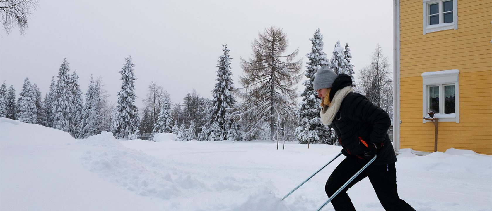 Nainen kolaa lunta keltaisen puutalon pihalla