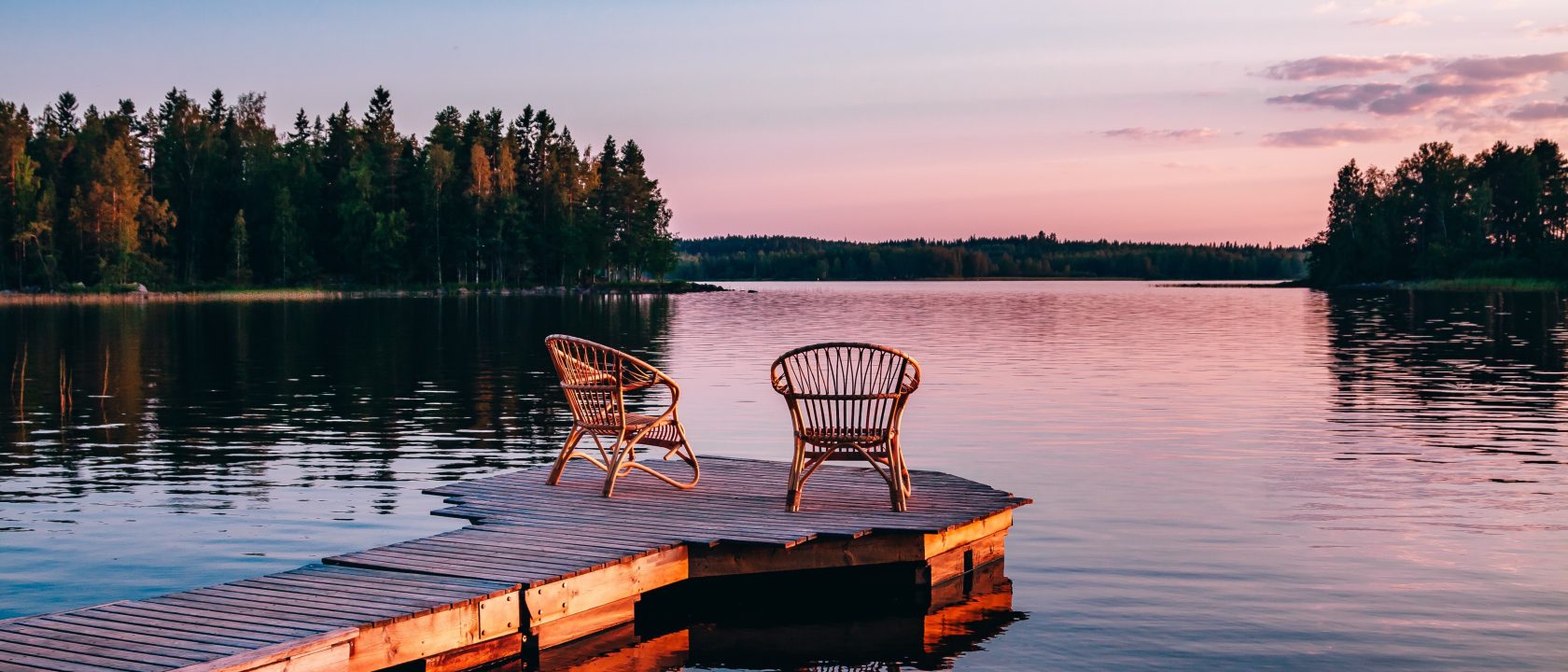 Kaksi rottinkituolia on aseteltu laiturille. Vesi on tyyni ja auringonlaskun väreissä hehkuva taivas heijastuu järven pinnasta.