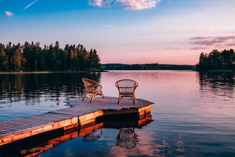 Kaksi rottinkituolia on aseteltu laiturille. Vesi on tyyni ja auringonlaskun väreissä hehkuva taivas heijastuu järven pinnasta.