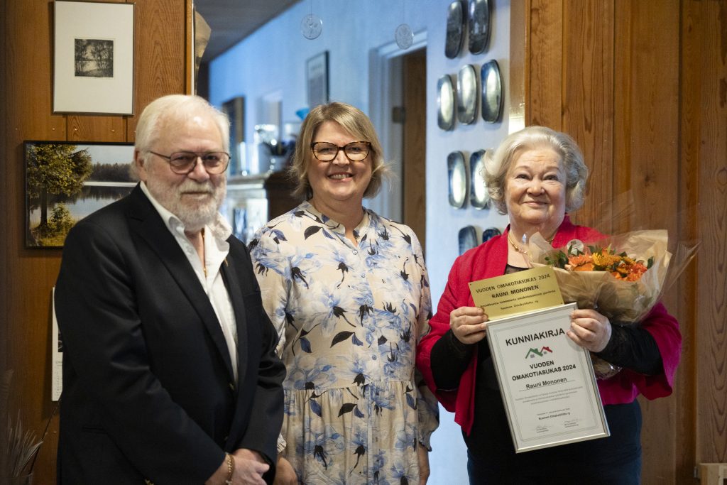 Kuvassa Omakotiliiton puheenjohtaja Ari Rehnfors, toiminnanjohtaja Marju Silander ja vuoden omakotiasukas Rauni Mononen, jolla sylissään kukat, kunniakirja ja onnittelulaatta.