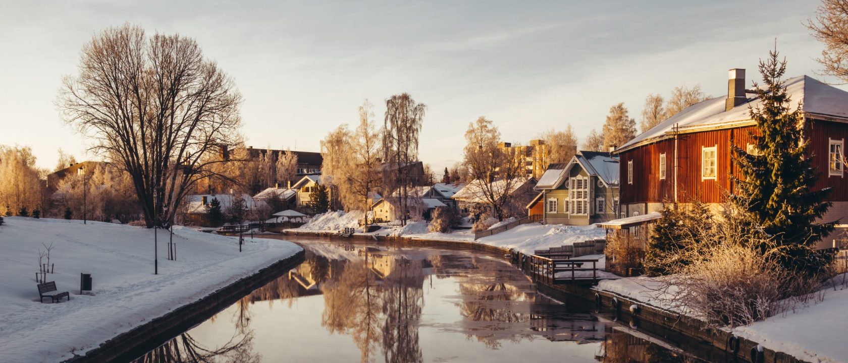 Talviaamun aurinko valaisee joen varressa olevia pientaloja, jotka heijastuvat tyynestä vedestä.