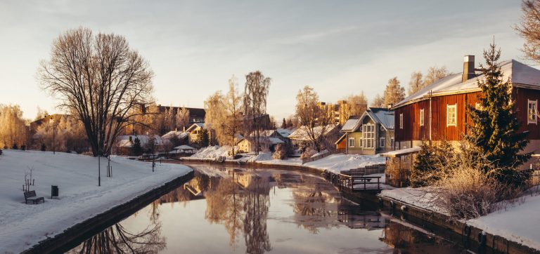 Talviaamun aurinko valaisee joen varressa olevia pientaloja, jotka heijastuvat tyynestä vedestä.